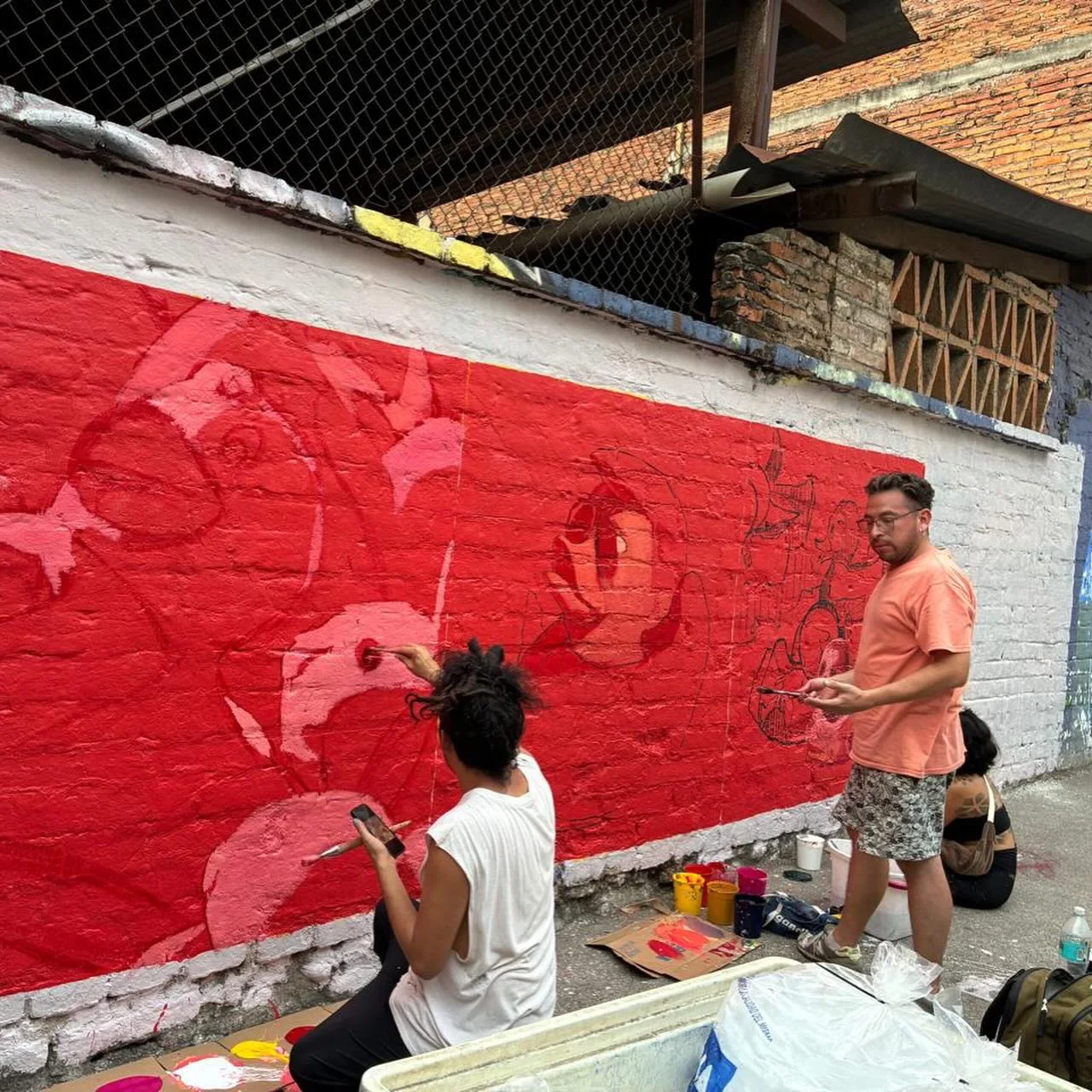 Two artists painting a red mural on a street wall, with paint pots on the ground