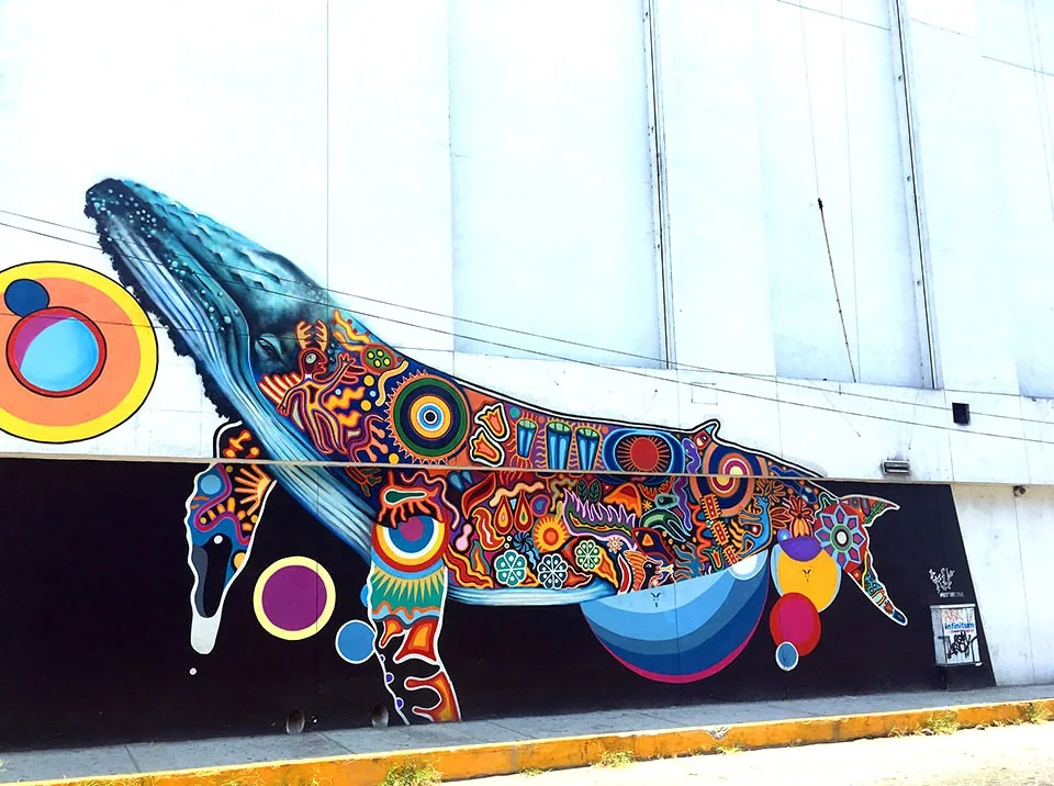 Large street mural of a humpback whale painted in vivid Mexican folk-art patterns